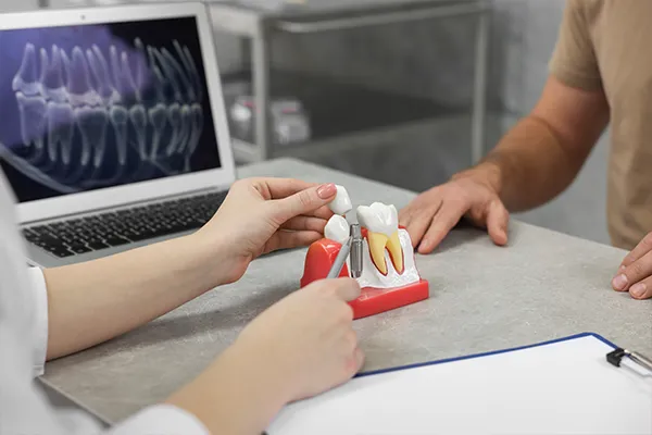 A dentist using a dental implant model to explain the procedure to a patient, with an X-ray displayed in the background.