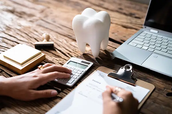 A man sitting at a desk with a calculator, laptop, and giant plastic tooth filling out insurance paperwork.